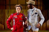 Two actors on stage, one dressed as a racing driver and the other wearing a striped blazer