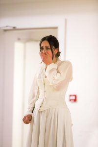 Actor dressed in white corset and white skirt clasping left hand to mouth