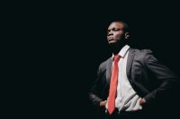 Nimshi Kongolo in suit with red tie in The Laramie Project