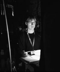 Black and white image of a stage manager, a man in black lothing lit by the desk light back stage