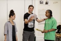 Three students rehearsing in a studio. The central student has their hand on one students shoulder and a script in the other hand