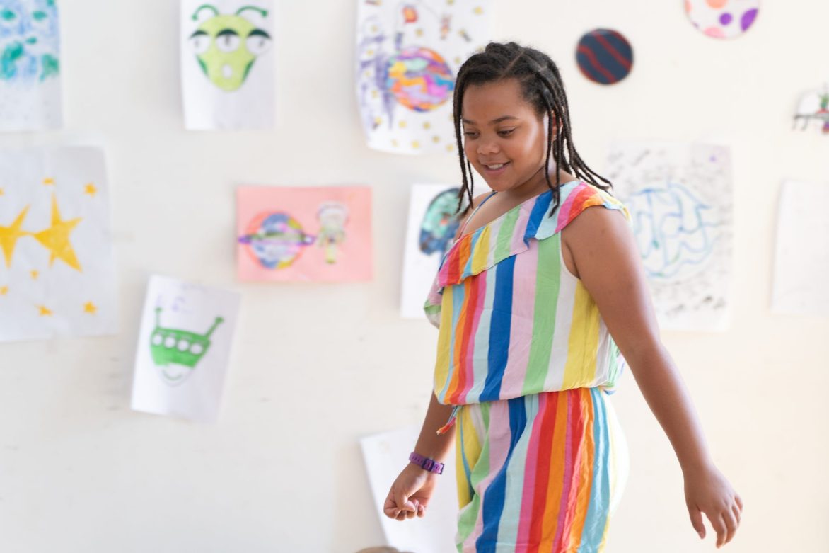 A member of the youth group is walking through a studio with costume designs on the wall behind
