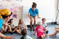 A youth group leader is entertaining a group of young artists who are sat on the floor around her. There are colourful drawings on the walls behind.